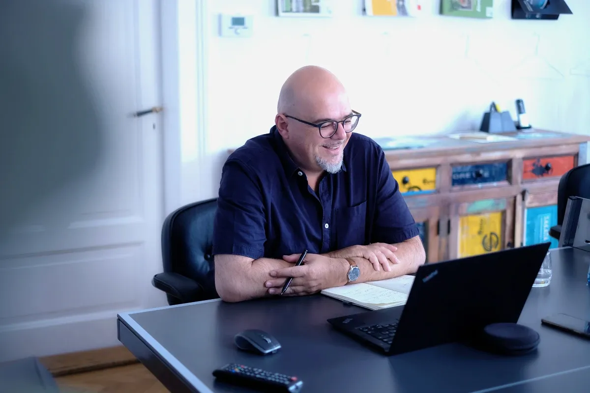 Boris Janda sitzt lächelnd am Schreibtisch vor einem aufgeklappten Laptop, daneben ein Notizbuch