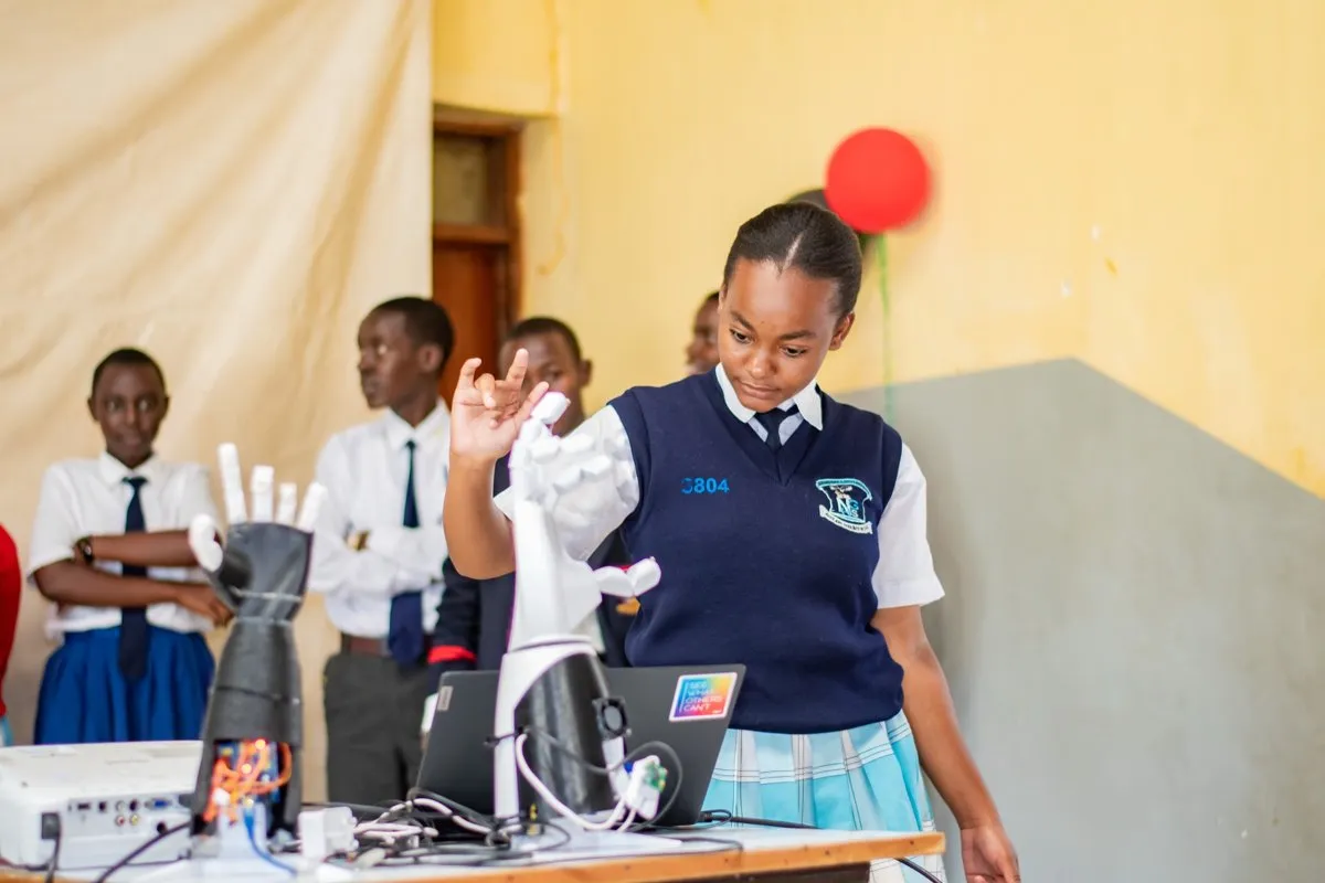 Eine Schülerin in Schuluniform hebt die Hand neben einer weißen Roboterhand auf einem Tisch, dahinter stehen weitere Schüler