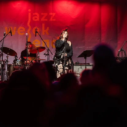 Jazzweekend © Dominik Hupf Über eine schattiges Publikum hinweg sieht man eine rot erleuchtete Bühne mit Sängerin und Schlagzeuger.